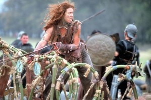A film still. Boudica, with wild red hair, rides a rough chariot into battle