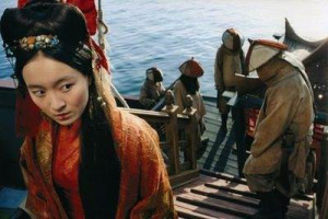 A still from Singing Behind Screens. The character of Ching Shih wears a red robe and an elaborate Chinese hairstyle. She glances behind her, to other sailors on her ship, dressed very plainly.