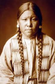A sepia-toned photograph of a Native American woman. She wears a loose shirt and a thick string of bone pipe beads as a mantle. She has long hair, put into two braids and silver medallion earrings