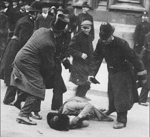 A black and white photo. A woman in Edwardina clothes lies (possibly unconscious) in the middle of the street. Several men and police offers are bent over her.