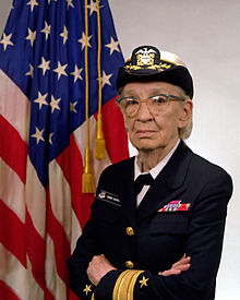An older woman in Naval dress blues and a white captain's cap. She stands with her arms crossed, an American flag behind her. 
