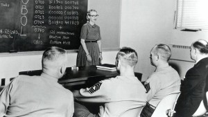 A black and white photo. A woman stands next to a chalkboard, instructing a group of men in military uniforms.