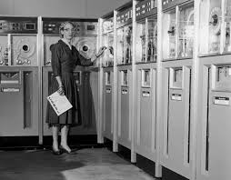 A black and white photo. A woman, wearing a 1950s style dress, holds some papers while looking at a bank of large computers.